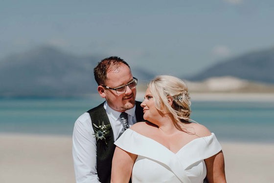 couple standing on beach with water behind them looking into each other's eyes in wedding dress and suit 